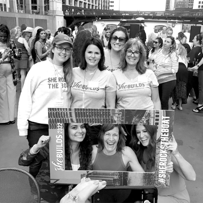 Black and white photo of a group of seven women posing with a frame that says 'She Builds 2022 #SheRocksTheBoat' with other women in the background.
