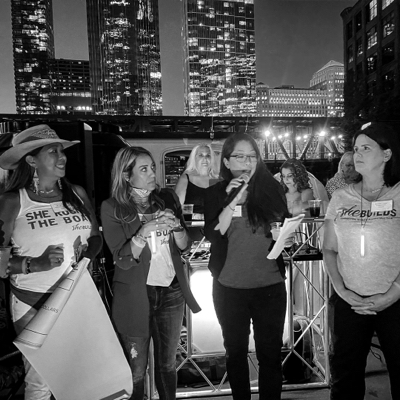 Black and white photo of a group of four women in front of nighttime Chicago skyline, one woman is speaking into a microphone.