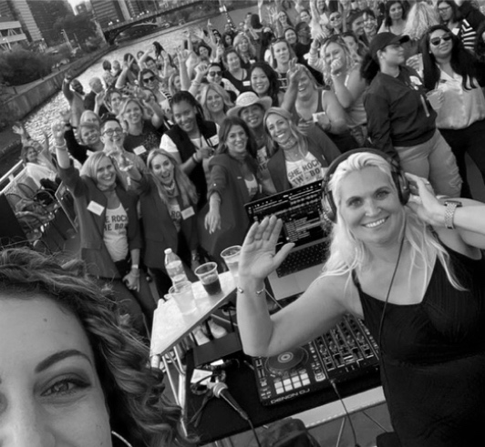 A woman DJ and attendees of a previous She Builds event waving at the camera while on a boat cruise.